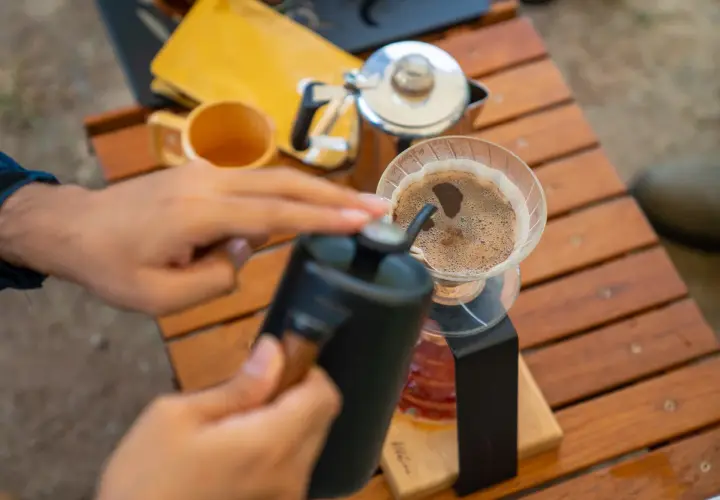 Ręce przygotowują aromatyczną kawę w górach. Na drewnianym stoliku stoi dripper z kawą, obok czajniczek i kubek.