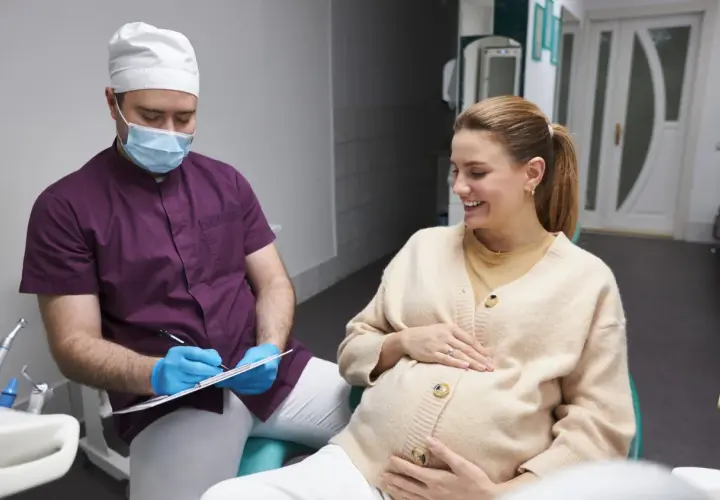 Dentista con mascherina parla con una donna incinta sorridente, discutendo della pulizia dei denti in gravidanza.