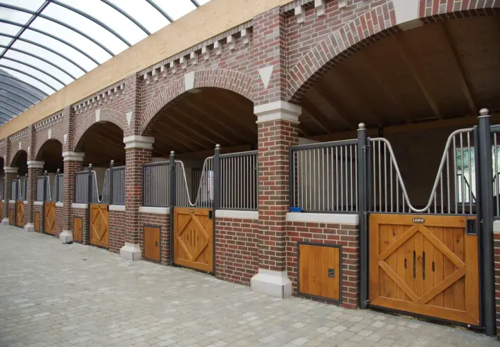 Rangée de stalles de chevaux en briques avec portes en bois et toiture transparente.