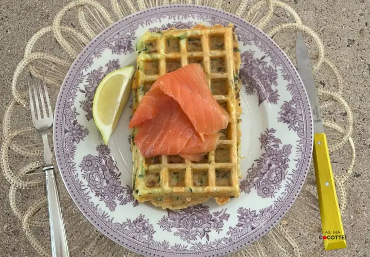 Gaufres salées garnies de saumon fumé et d'un quartier de citron, sur une assiette ornée, avec une fourchette et un couteau à manche jaune.
