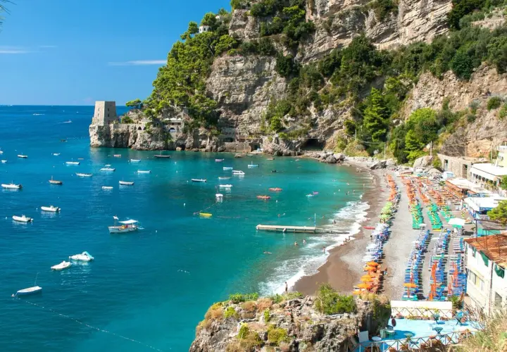 Vue panoramique d'une plage de galets animée à Almafi, Italie, avec des parasols colorés, des chaises longues et des eaux bleues claires parsemées de bateaux.