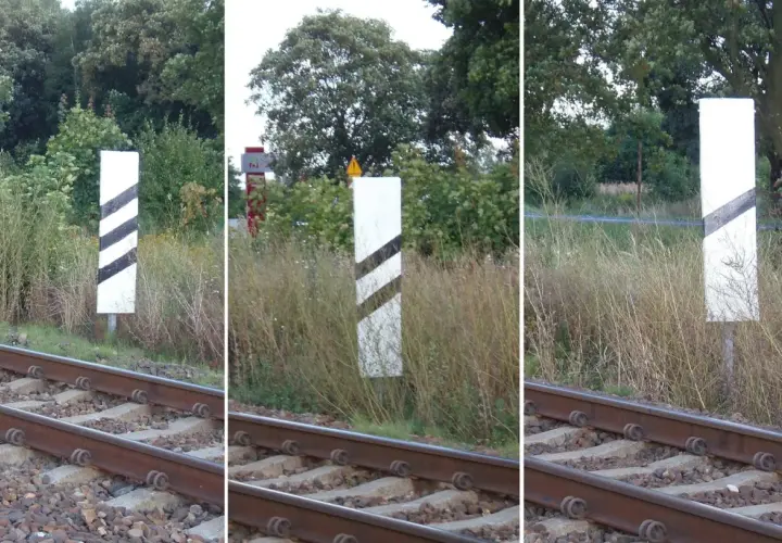 Trzy białe słupki z czarnymi pasami, wskaźnik w11a, stoją przy torach kolejowych.