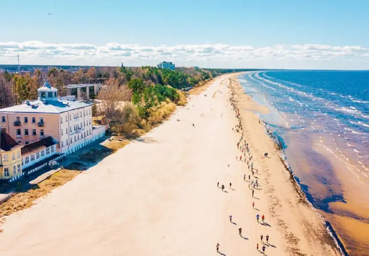 Szeroka, piaszczysta plaża na Łotwie, z budynkami przy brzegu i ludźmi spacerującymi wzdłuż morza.