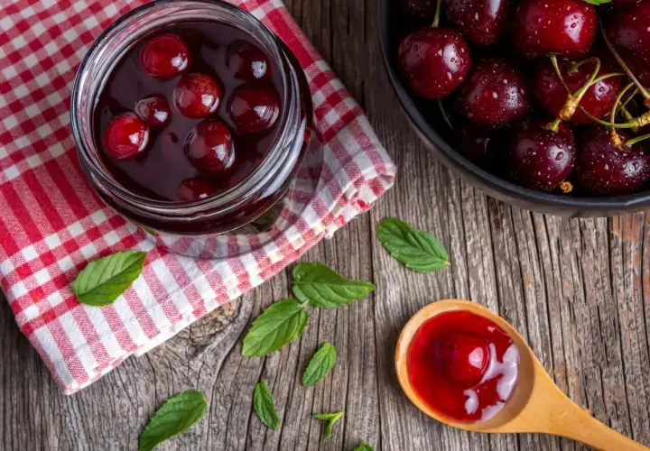 Słoik pełen domowego dżemu z czereśni, obok miska świeżych owoców i drewniana łyżka z porcją słodkiego przysmaku.