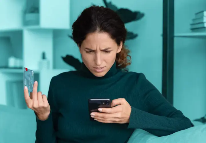Zmarszczona kobieta trzyma kartę i telefon, zastanawiając się nad bezpieczne zakupy w internecie.