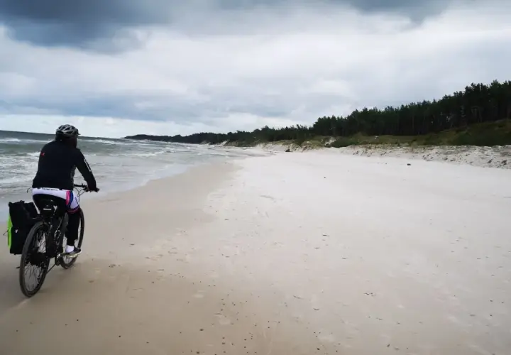 Rowerzystka przemierza piaszczystą plażę na mierzei sarbskiej, z widokiem na wzburzone morze i sosnowy las.