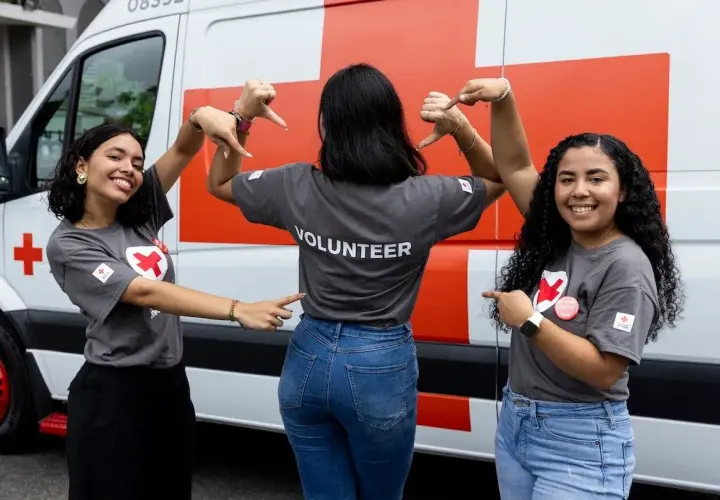 Three young women in "VOLUNTEER" shirts pose in front of a Red Cross van, smiling and pointing.