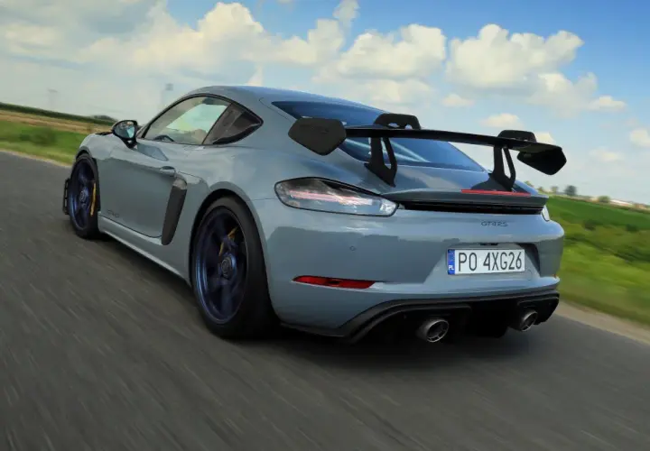 Szary Porsche GT4 RS z dużym skrzydłem pędzi po drodze, zostawiając za sobą rozmyte pola i błękitne niebo.