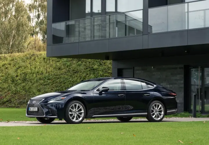 Elegancki, ciemnogranatowy Lexus LS 500 zaparkowany przed nowoczesnym domem z dużymi oknami i zielonym ogrodem.