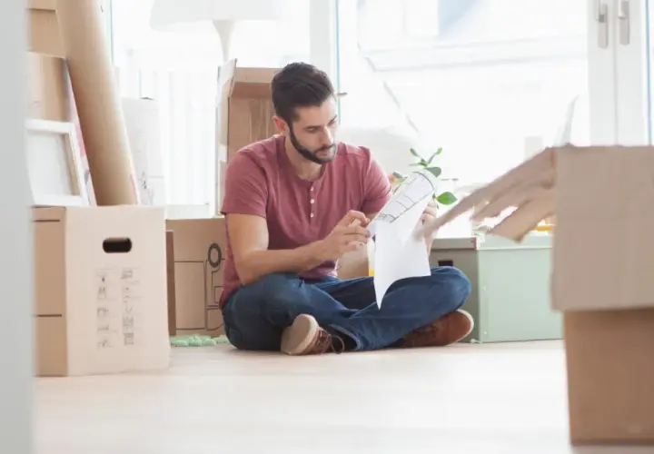 Hombre revisa un plano rodeado de cajas, preparándose para la mudanza. Podría ser el **contrato compra venta** de su nuevo hogar.