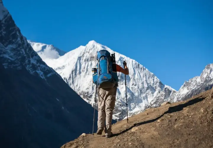 Un excursionista con mochila azul asciende un sendero polvoriento, bastones en mano, ante majestuosas montañas nevadas. Una aventura de trekking Manaslu.