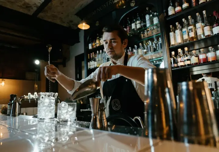 Barman przygotowuje drinka, zastanawiając się, ile zarabia dobry barman. Za nim półki z alkoholem.
