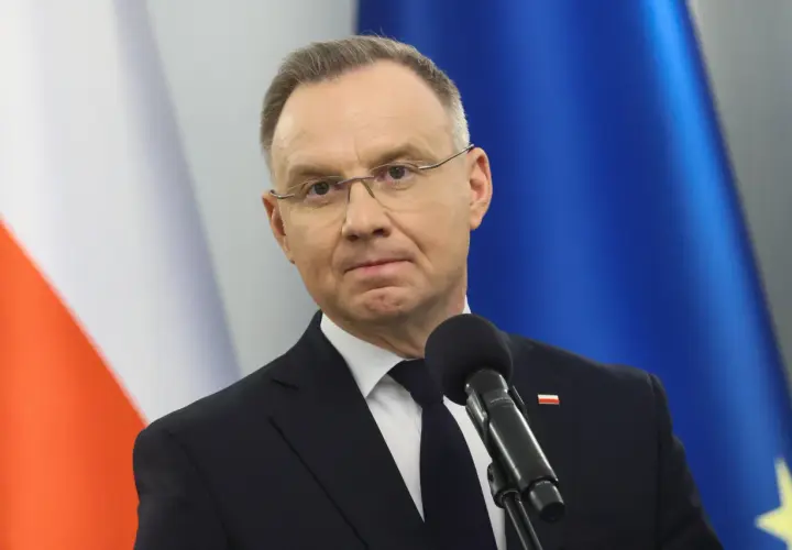 Prezydent Andrzej Duda w garniturze przed mikrofonem, w tle flagi Polski i UE.