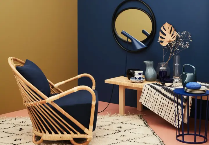 Ein Rattan-Sessel mit dunkelblauem Kissen steht auf einem flauschigen Teppich. Welche Farbe passt zu Gold? Hier: ein Spiegel mit goldenem Rahmen und ein gelber Wandbereich.