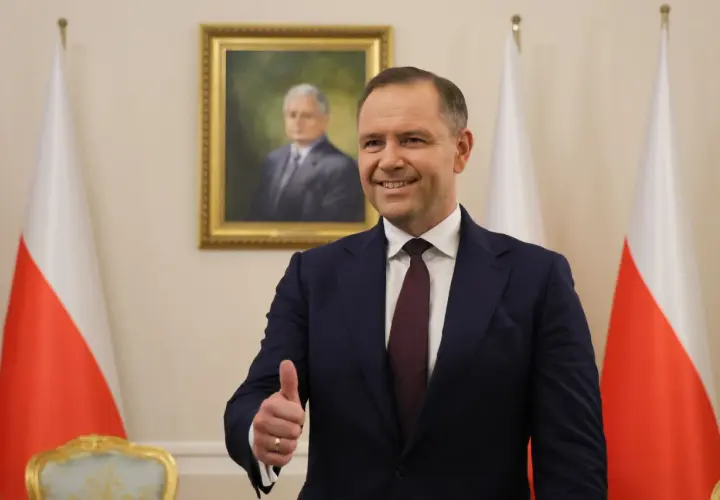 Mężczyzna w garniturze pokazuje kciuk w górę na tle portretu i flag Polski.