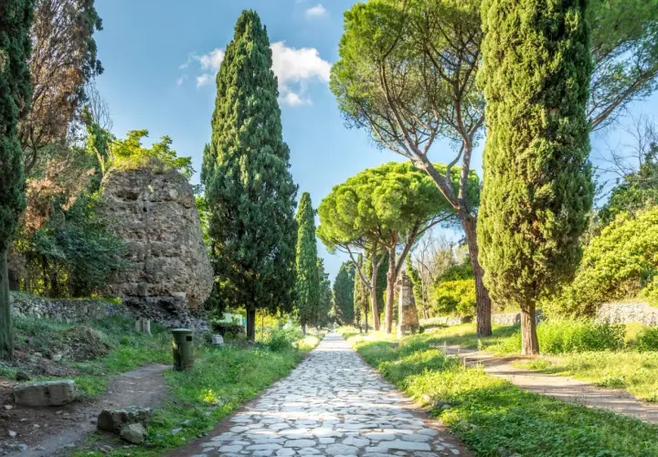 Allée pavée bordée de cyprès et de pins sur la Via Appia, avec des ruines antiques sur le côté.