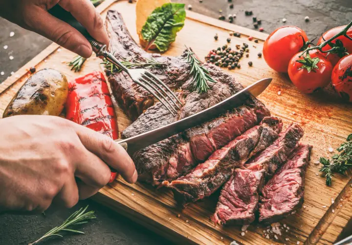 Une main coupe un succulent côte de bœuf barbecue sur une planche en bois, accompagné de pommes de terre rôties et de poivrons grillés.