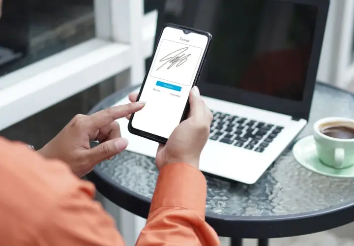 Persona usando su móvil para firmar online documento. En la mesa hay un portátil y una taza de café.