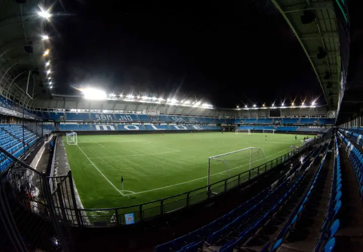 Piłkarski stadion Molde nocą, oświetlony jaskrawymi światłami, z pustymi niebieskimi trybunami i zieloną murawą.