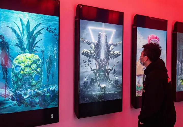 Ein Mann betrachtet digitale Kunstwerke, die an einer Wand hängen. Die Werke zeigen surreale Szenen, wie eine Ananas unter Wasser mit Skeletten und eine symmetrische Figur mit vielen Armen. Diese Werke stammen von einem berühmten Grafik Künstler.