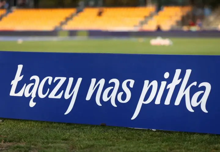 Niebieska tablica z napisem "łączy nas piłka" na tle stadionu. To motto, które jednoczy kibiców i prezesa PZPN.