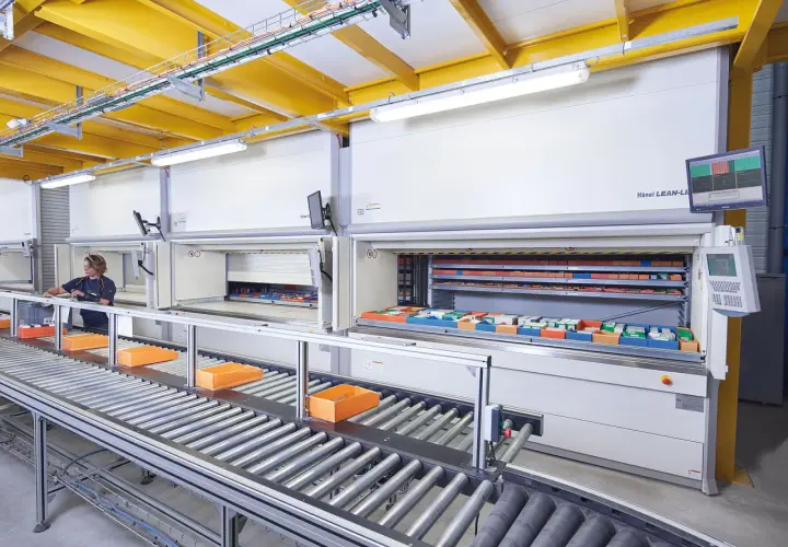 Ein Kommissionierlager mit automatisierten Lagersystemen. Eine Mitarbeiterin entnimmt Teile, während Behälter auf einem Rollenband transportiert werden.