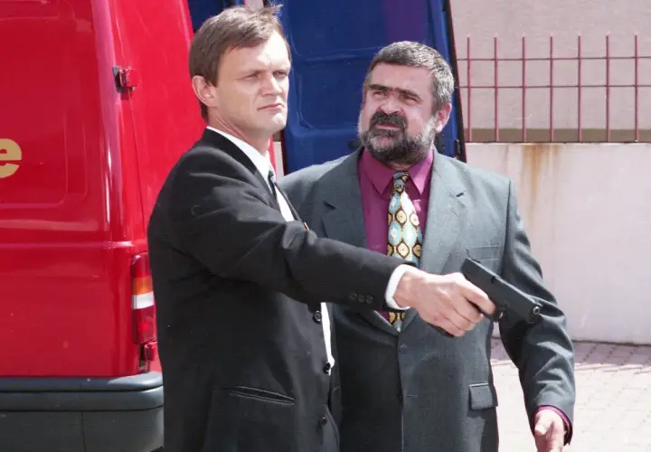 Gangster z filmu Kiler, w garniturze, celuje z pistoletu. Obok stoi mężczyzna w czarnym garniturze.