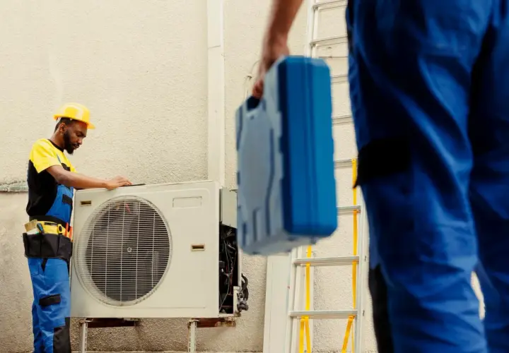 Técnicos revisan unidad exterior de aire acondicionado que dejó de funcionar de repente. Uno con casco amarillo examina la máquina, otro lleva una caja de herramientas azul.