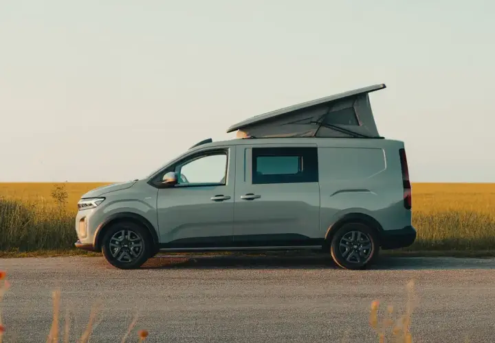 Un Dacia Sandman gris, prêt pour l'aventure, avec son toit relevable ouvert, stationné au bord d'un champ doré au coucher du soleil.