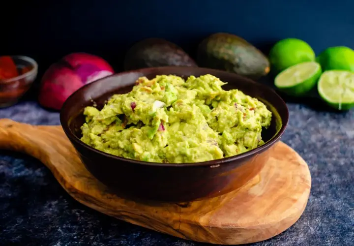 Miseczka guacamole z kawałkami cebuli i pomidorów, na drewnianej desce, w tle awokado i limonki.