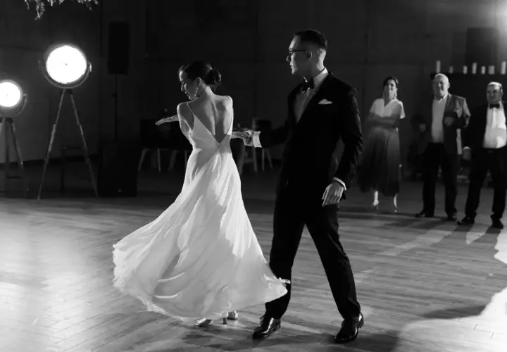 Para młoda tańczy, co zamiast pierwszego tańca. Ona w zwiewnej sukni, on w eleganckim garniturze. W tle goście.