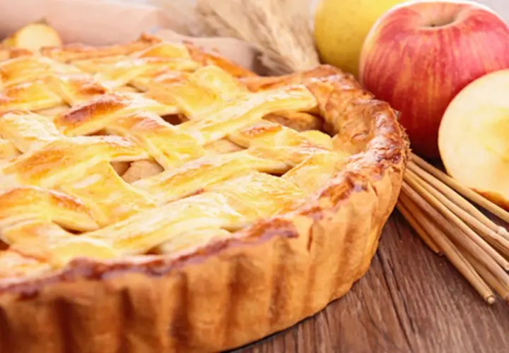 Tarta de manzana dorada con rejilla de masa, junto a manzanas frescas y palitos de canela.