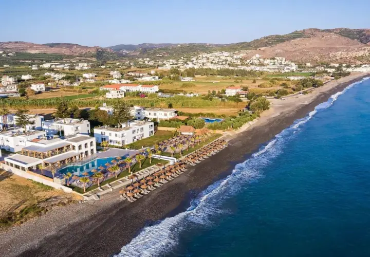 Hotel Solimar White Pearl, białe budynki z basenem i leżakami, rozciągają się wzdłuż ciemnej plaży i turkusowego morza.