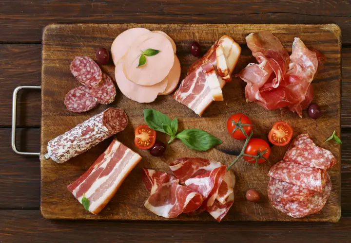 Assortiment de différents types de charcuterie : saucisson, jambon cru, bacon, mortadelle, accompagnés de tomates cerises et d'olives.