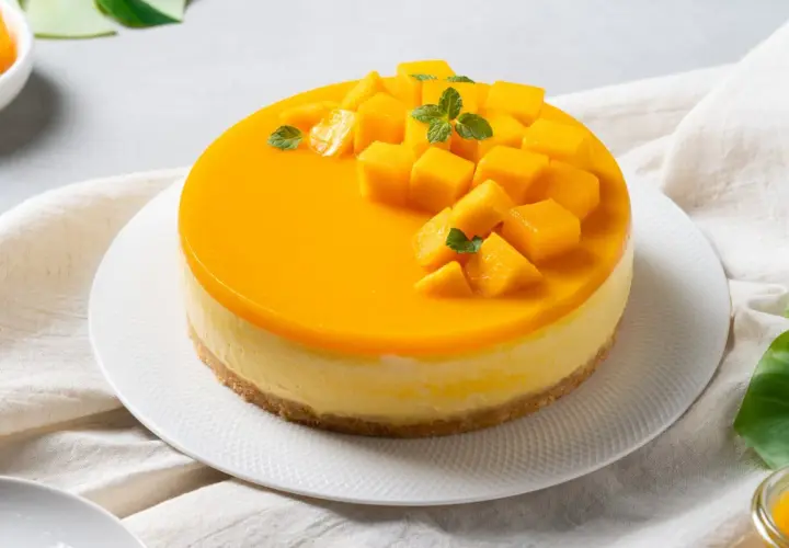 Sernik z błyszczącą polewą mango, ozdobiony kostkami mango i listkami mięty.
