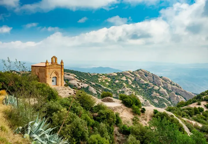 Pequeña ermita en la cima de una colina, con vistas a las impresionantes rutas de Montserrat y un paisaje montañoso bajo un cielo azul con nubes.