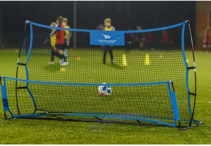 Piłkarski rebounder Yakimasport na zielonej murawie, z piłką w siatce. W tle trening młodych piłkarzy.