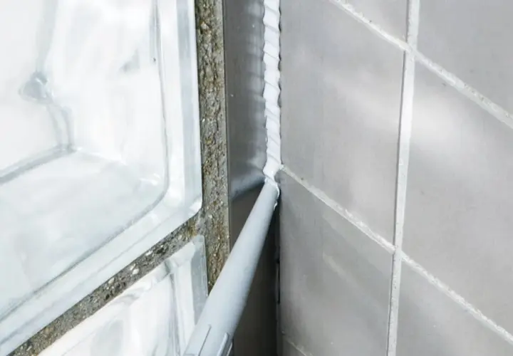 Applicator sprutar vit silikonfog längs en fog i badrummet, intill en duschvägg av glasblock.