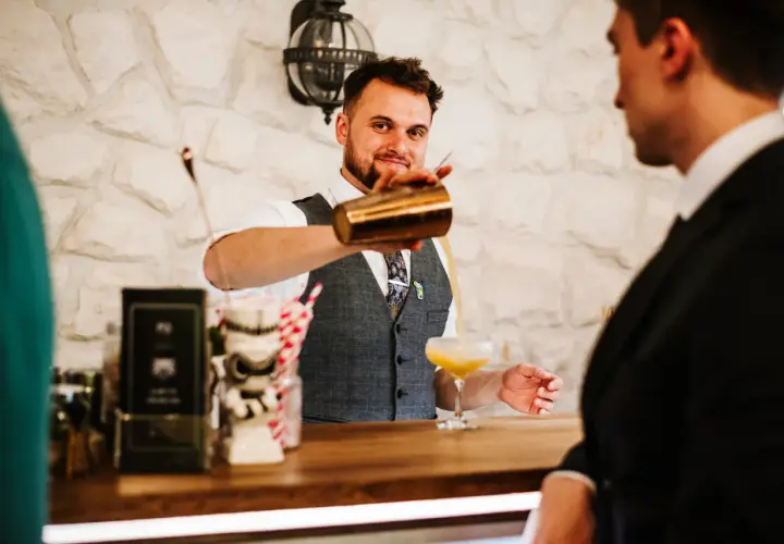 Uśmiechnięty barman w kamizelce nalewa drinka do kieliszka.