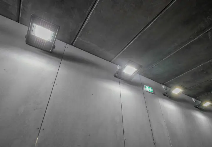 Jasne, oto opis alternatywny:

Oświetlenie długiego korytarza z betonowym sufitem i szarymi ścianami. Kilka nowoczesnych lamp LED rzuca jasne światło, a zielony znak ewakuacyjny wskazuje drogę.