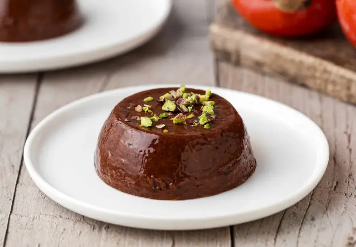 Budino di cachi al cioccolato, guarnito con pistacchi tritati, servito su un piatto bianco.