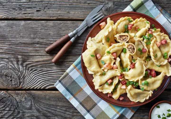 Pyszne pierogi z mięsem, posypane skwarkami i szczypiorkiem, czekają na talerzu. Obok sztućce i miseczka śmietany.