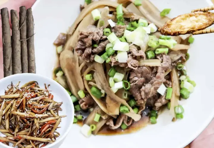 A delicious dish of braised burdock with thinly sliced beef and green onions, served on a white plate. A small bowl of burdock stir-fry is on the side.