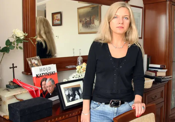 Kobieta z blond włosami w czarnej bluzce i jeansach, obok książka "Hołd Katyński" i ramka ze zdjęciem.