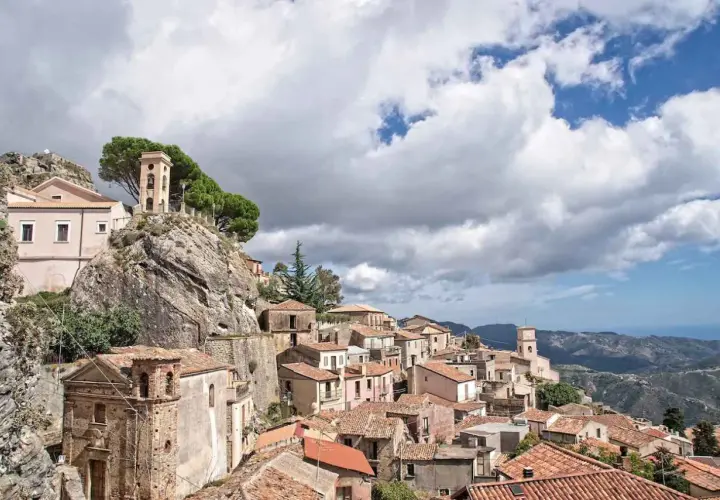 Charming towns in Calabria, Italy, cling to a rocky hillside under a dramatic sky. A bell tower stands sentinel over terracotta roofs.