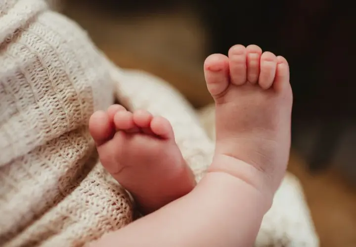 Delikatne, różowe stópki niemowlaka z lekko podwiniętymi palcami u nóg, otulone miękkim, beżowym kocykiem.