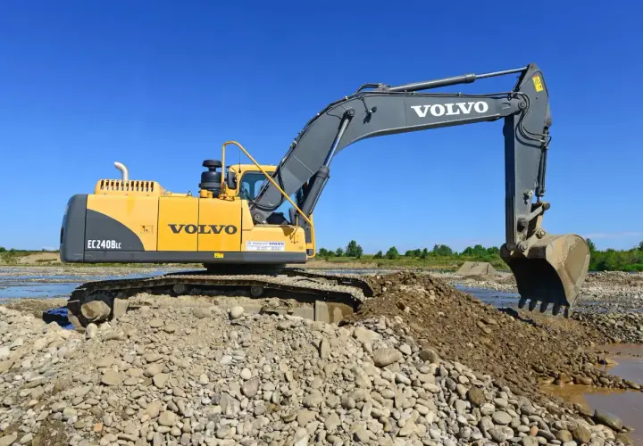 Żółta koparka Volvo EC240BLc pracuje na kamienistym brzegu rzeki. Zastanawiasz się, ile bierze koparka za godzinę?