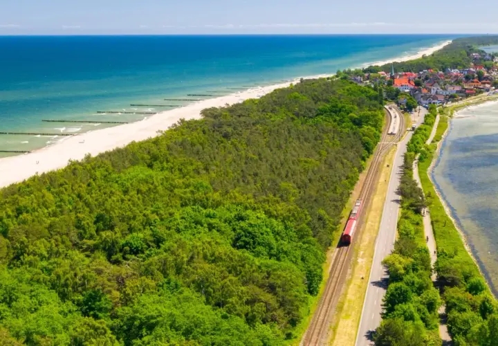 Czerwony pociąg jedzie wzdłuż wybrzeża. W tle widać plażę, morze i nadmorską miejscowość. Idealna podróż pociągiem nad morze.