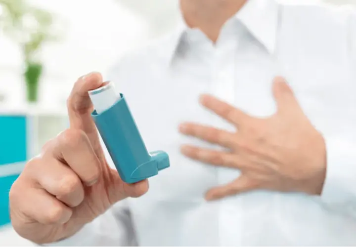 Mann hält Inhalator mit ipratropiumbromid. Seine andere Hand liegt auf der Brust, was auf Atemnot hindeutet.