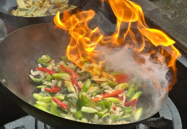 Ogień płonie nad warzywami w woku, tworząc aromatyczny stir fry. W tle widać smażone ziemniaki.